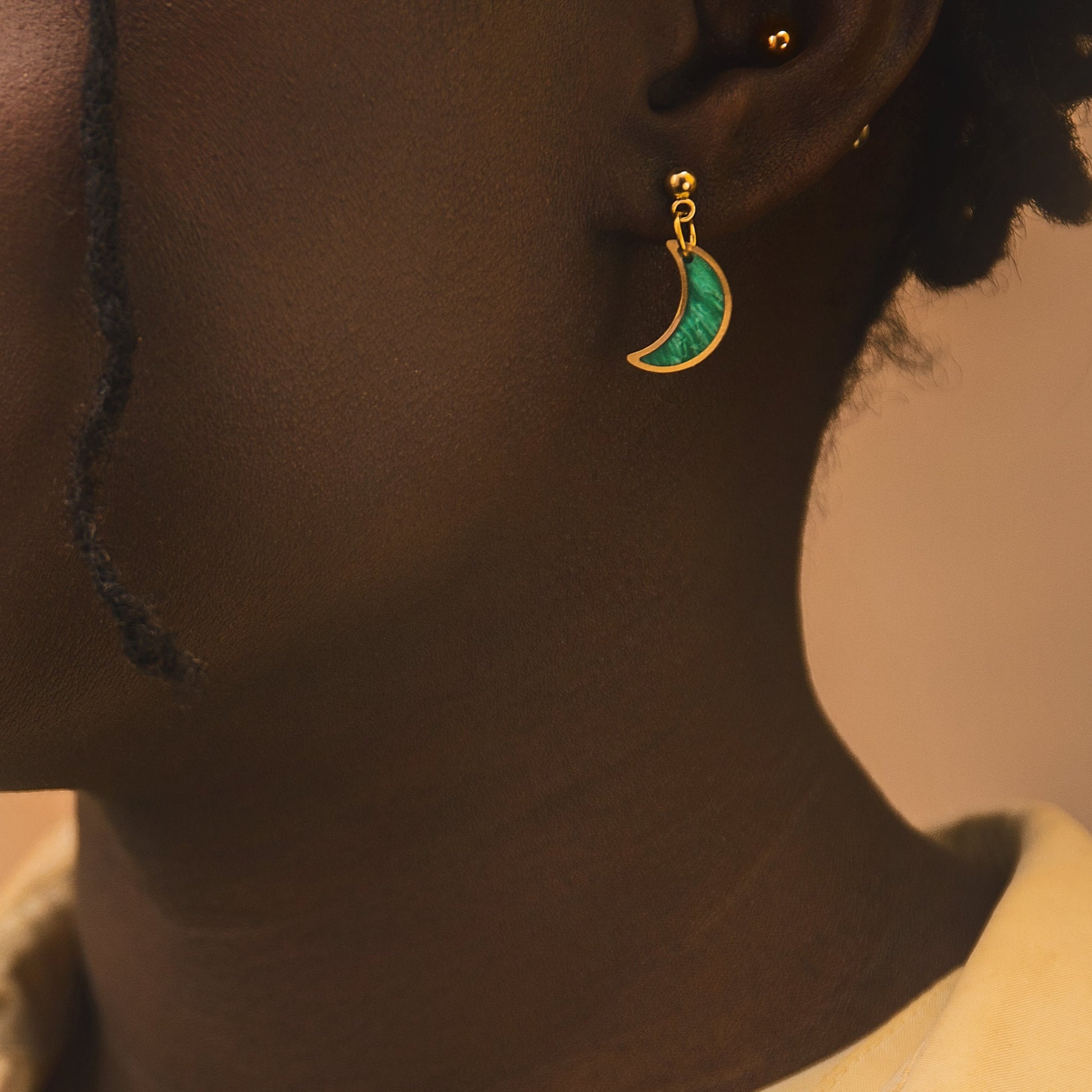 Close-up of a person wearing a yellow shirt and a green earring with a crescent moon design on a beige background.