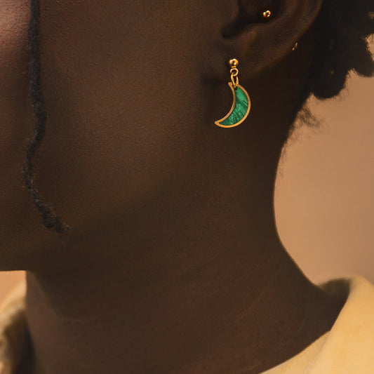 Close-up of a person wearing a yellow shirt and a green earring with a crescent moon design on a beige background.